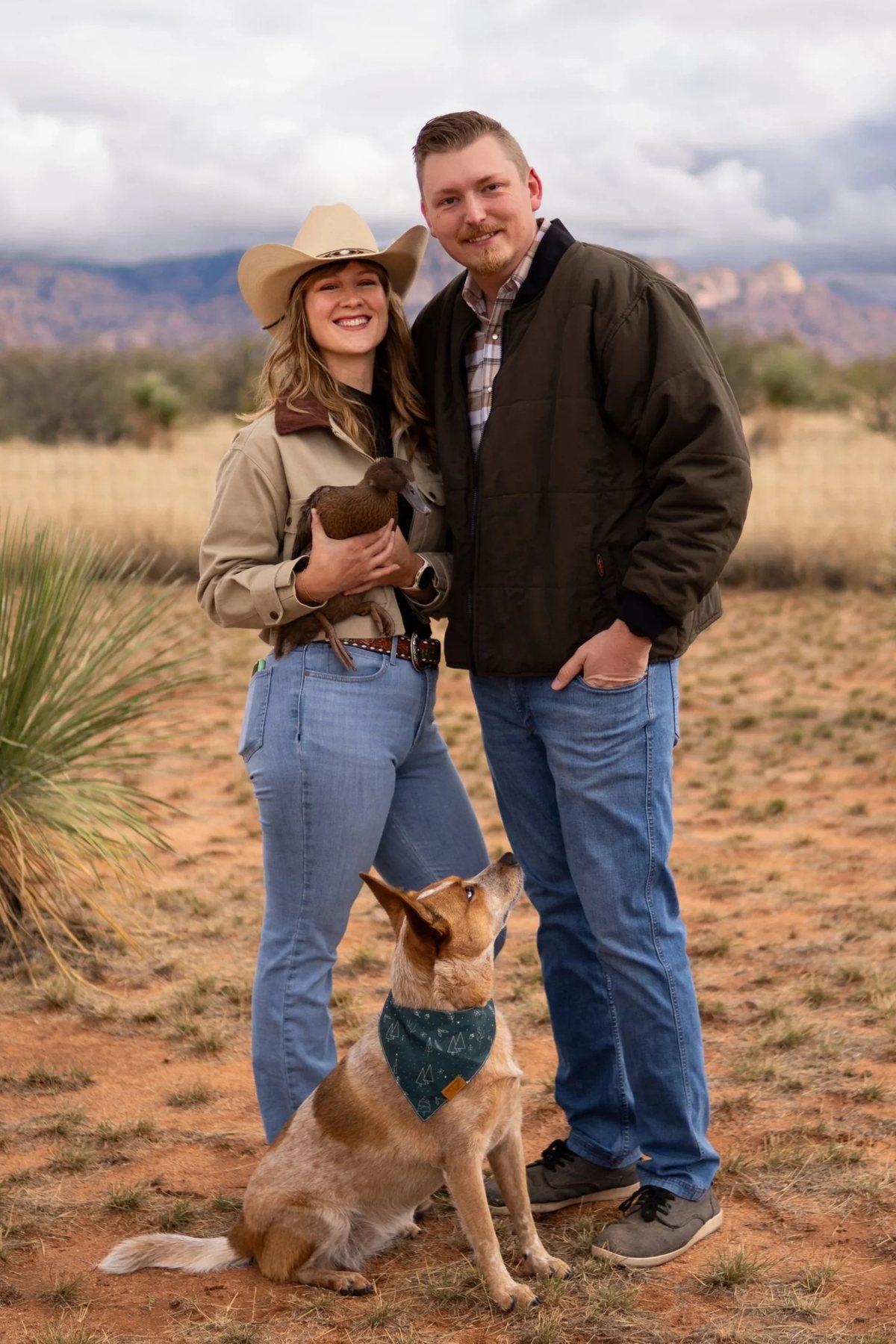 James and Kaylin Roth outdoors in the Arizona desert with their dog and duck, mountains behind them.